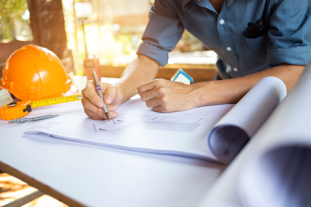 A person uses a pen to design ductwork on a blueprint with an orange construction hat and measuring tape on the same table.