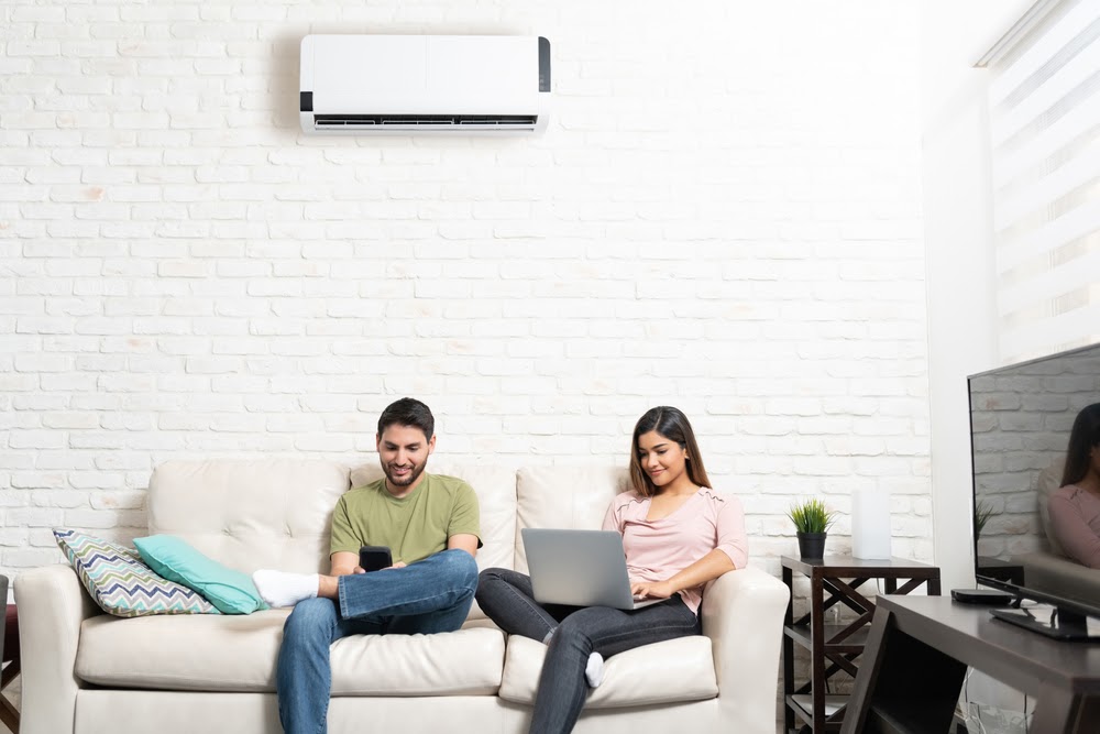 A man and woman sitting on a couch with a mini split system on the wall behind them.