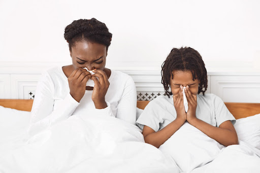 A mom and child sitting in bed and blowing their noses.